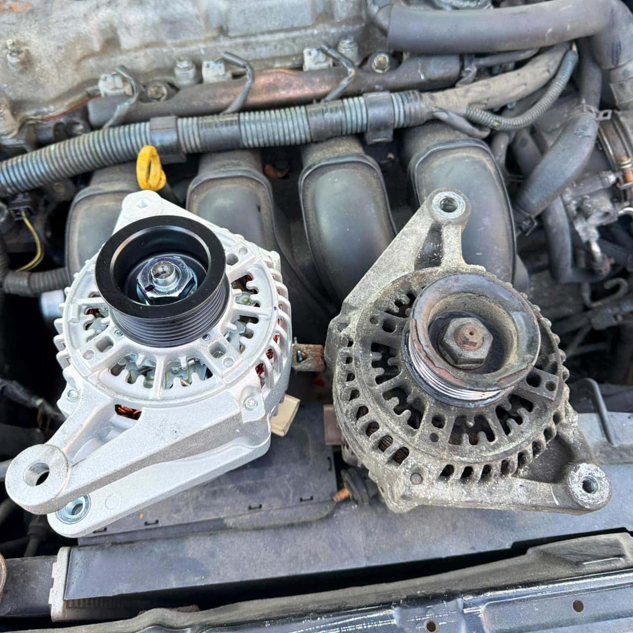 A brand-new alternator next to the old corroded unit it replaced, over the engine bay