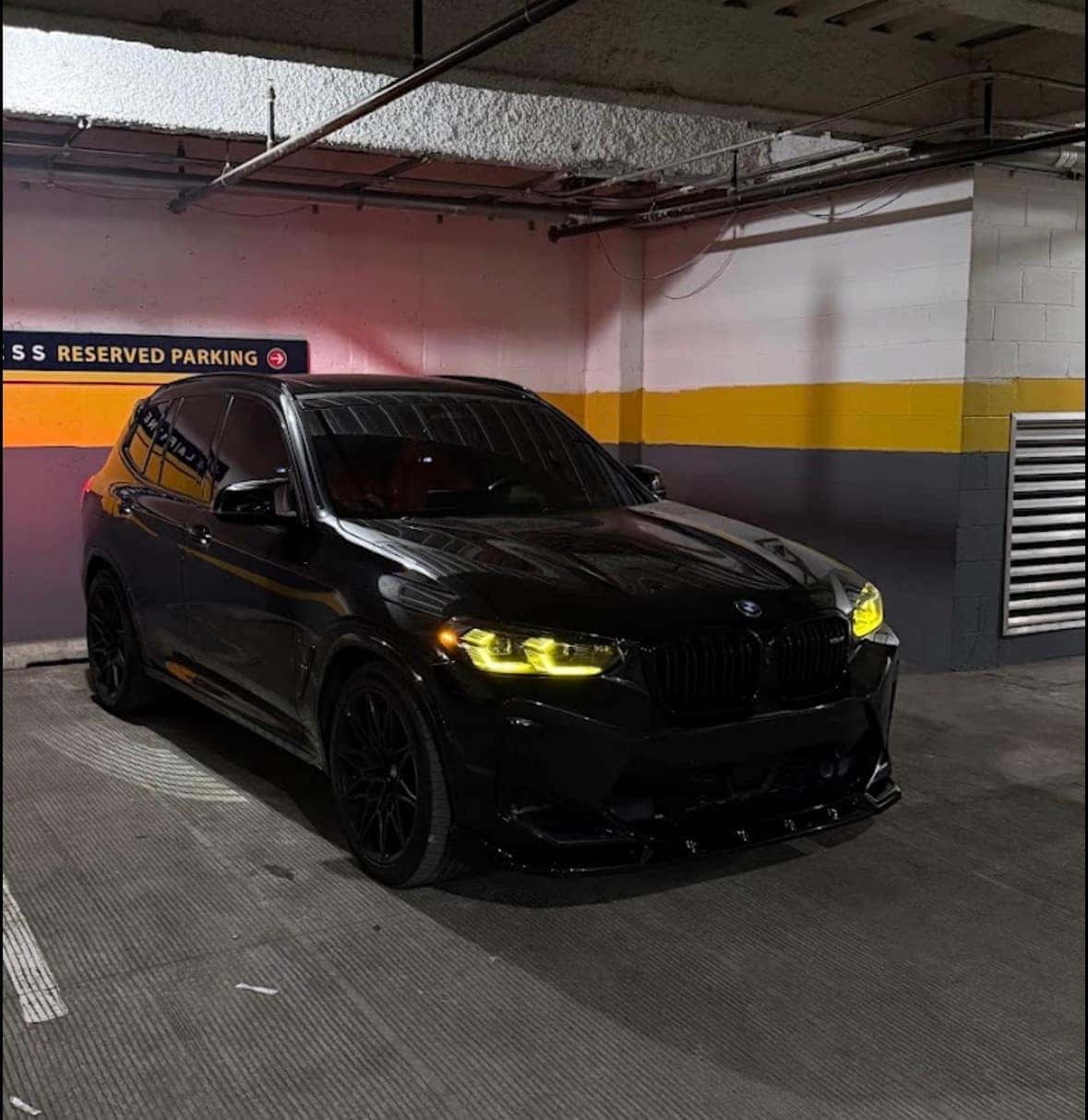 Blacked-out BMW X3 M with yellow laser headlights and gloss-black wheels in a parking garage